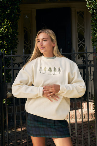 A woman with long blonde hair wears the Christmas Tree Farm Embroidered Crewneck and a plaid skirt, standing in front of a black iron gate and house. She smiles slightly with her hands crossed.