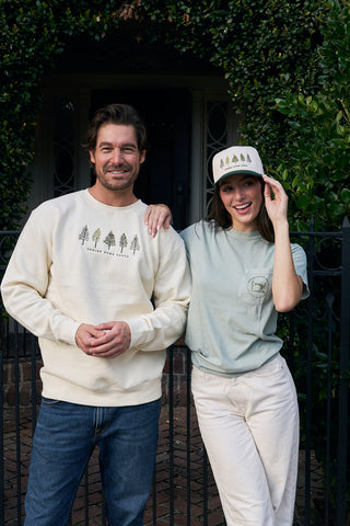 A man in a cream sweatshirt with tree graphics and a woman in a green t-shirt and the Christmas Tree Farm Embroidered Hat stand smiling together outside a house, surrounded by greenery and a black gate.