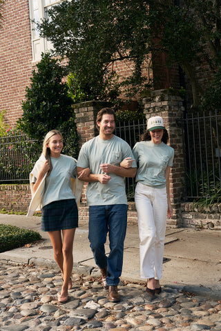 Three friends stroll arm-in-arm on a cobblestone street, each wearing a light green Christmas Tree Farm Tee. The man in the center is flanked by two women—one in a skirt, the other in white pants and a hat, with trees and a brick building behind them.