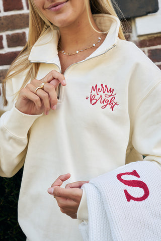 A woman wearing the cream Merry & Bright Quarter Zip—an embroidered sweatshirt—holds a white knit blanket with a bold red “S” and smiles in front of a classic brick wall.