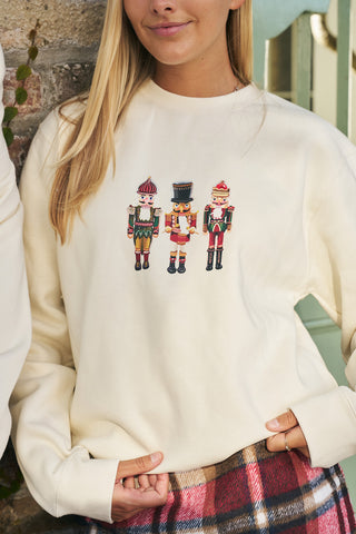 A woman stands by a stone wall and stair railing, wearing the Nutcracker Crewneck Sweatshirt with three colorful nutcracker designs, paired with a plaid skirt.