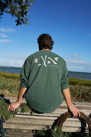 A man with brown hair sits on a wooden bench by the water, surrounded by greenery, wearing a green Winter Sports Gear Long Sleeve Tee. The sky above is blue with clouds.
