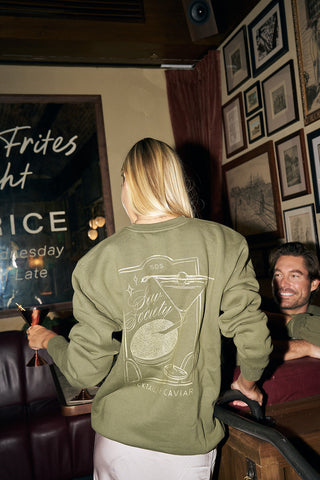 A woman with long blonde hair, wearing a Cocktails & Caviar Crewneck Sweatshirt, stands in a cozy, art-filled bar. A smiling man with a drink sits nearby, enjoying the inviting vibe of this festive lounge wear scene.