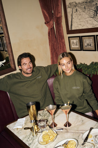 A man and woman, both wearing matching olive green Cocktails & Caviar Crewneck Sweatshirts, smile as they enjoy cocktails and chips together at a marble table in a restaurant booth, with framed pictures on the wall behind them.