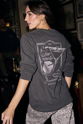 A woman with long brown hair wears the Cocktails & Caviar Long Sleeve Tee, a festive unisex top perfect for your next holiday party. It features a martini glass and the phrase "SDS, Stay Local, Cocktails Always" on the back.