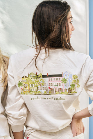 A woman with long brown hair wears the Holiday Rainbow Row Long Sleeve Tee, which displays colorful houses and trees with "Charleston, South Carolina" text on the back. She is turned to the side, showing off this festive design.