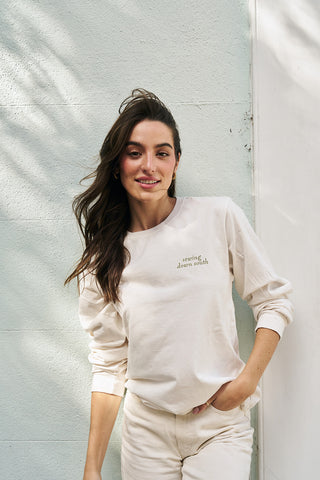 A woman with long brown hair smiles as she leans against a pale wall, wearing the Holiday Rainbow Row Long Sleeve Tee and matching pants, sunlight casting soft shadows on the wall behind her.