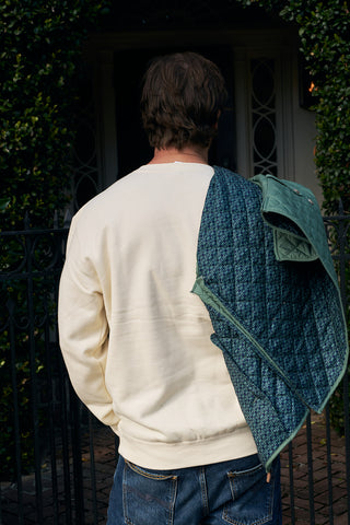 A person with brown hair, wearing the Christmas Tree Farm Embroidered Crewneck and blue jeans, stands with a green quilted jacket over one shoulder in front of a black iron gate surrounded by foliage, facing away from the camera.
