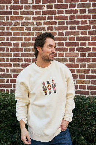 A man wearing a cream Nutcracker Crewneck Sweatshirt featuring three nutcrackers on the front stands by a brick wall, looking to the side with one hand in his jeans pocket.