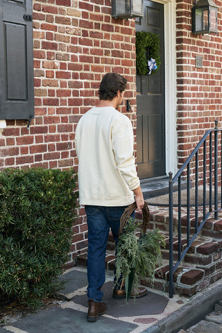 A man in a Nutcracker Crewneck Sweatshirt and jeans stands on a stone path, holding greenery and a wreath, about to climb steps to a brick house with a dark door decorated with a wreath.
