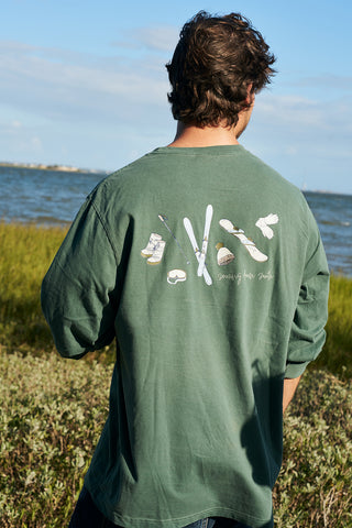 A person with wavy brown hair wearing a green Winter Sports Gear Long Sleeve Tee stands outdoors by the water. The back showcases illustrated fishing gear and the phrase “Coming in from Smoke.”.