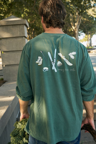 A person with brown hair stands outdoors holding leafy greens, wearing the Winter Sports Gear Long Sleeve Tee in green, featuring winter sports illustrations, surrounded by trees and concrete structures.