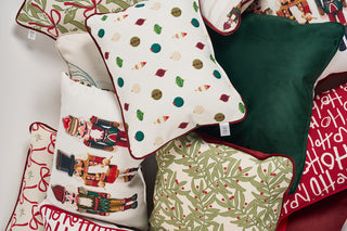 A pile of festive holiday pillows featuring patterns like nutcrackers, holly leaves, ornaments, candy canes, green velvet, and red text reading “Happy Holidays” on a white background.