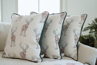 Three Repeating Reindeer Pillows featuring a white background and illustrated reindeer designs are arranged on a light-colored couch in a bright room with a window behind them.