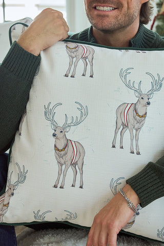 A person in a green sweater smiles while holding the Repeating Reindeer Pillow, which features festive red and gold reindeer harnesses. Only their lower face and arms are visible, putting the charming design in focus.