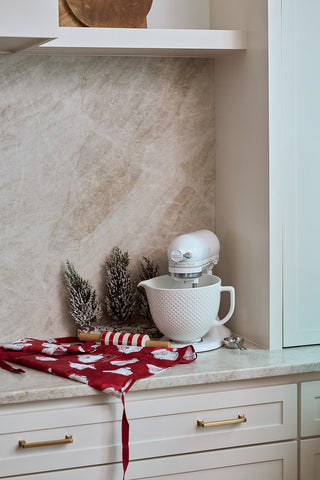 A white stand mixer sits on a kitchen counter next to small pine trees, a Santa Apron with adjustable neck strap, a striped rolling pin, and a metal measuring spoon, all set against a light marble backsplash.