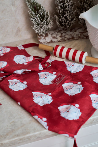 The Santa Apron, featuring an adjustable neck strap, rests on the counter beside a red and white striped rolling pin, with snow-dusted pine trees and a white mixing bowl adding to this festive Sewing Down South scene.
