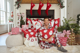 A man in plaid pajamas smiles on a fluffy rug before a fireplace decorated for Christmas with stockings labeled “SDS,” a Vintage Repeating Santa Pillow, wrapped gifts, and greenery.
