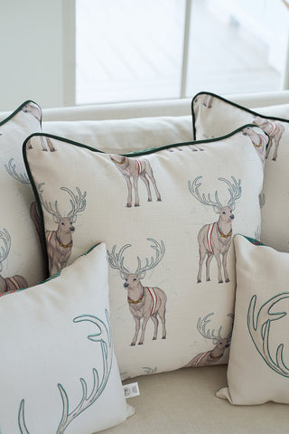 A group of holiday pillows on a light sofa includes the Repeating Reindeer Pillow, which features red harnesses. Smaller pillows with antler designs and a green velvet accent pillow complete the look as sunlight streams in from behind.