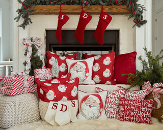 A festive fireplace decorated with red stockings, a Handwritten Ho Ho Ho Pillow, and a Vintage Stitch Santa Pillow. Wrapped gifts and greenery surround the hearth, creating a cozy Christmas scene.
