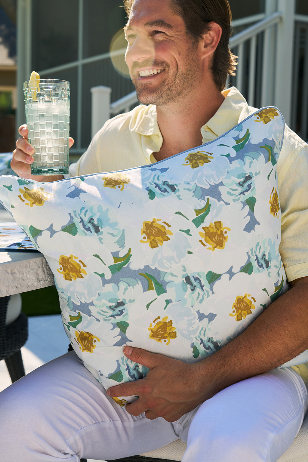 A man in a yellow shirt and white pants sits on a patio chair, drink in hand, smiling and relaxed. Nearby is the Blossoming Peonies Oversized Outdoor Pillow. The scene is outdoor with a table, stairs, and house in the backdrop.