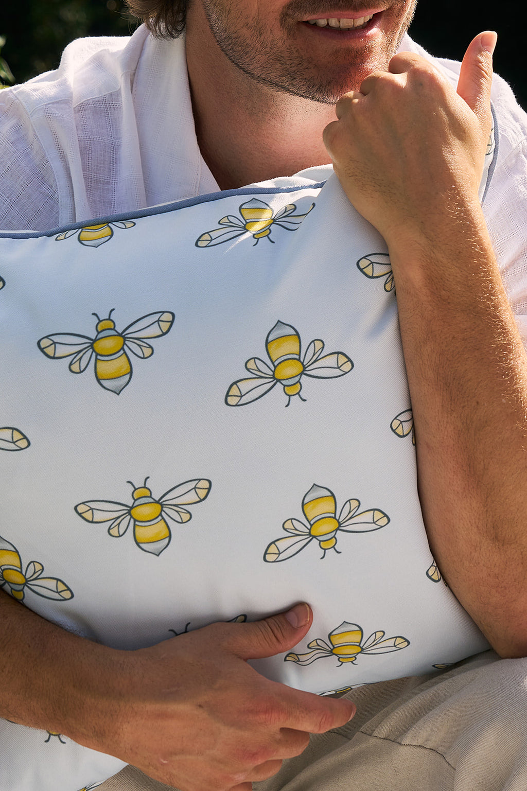 An outdoor lounge area features a beige chaise longue with light blue cushions adorned with Sewing Down Souths yellow and black bee patterns. Nearby, a basket holds matching Bee Trio Outdoor Pillows against lush greenery and colorful flowers in sunlight.
