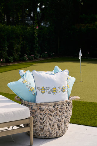 A woven basket with Bee Trio Repeating Bees Outdoor Pillows from Sewing Down South sits on a patio, displaying intricate designs. Nearby, a putting green with a white flag enhances the serene area, surrounded by lush greenery that boosts the tranquil atmosphere.