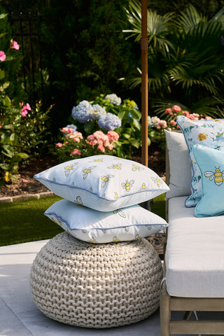 An outdoor patio showcases a beige knit pouf with two white Bee Trio with Repeating Bees Outdoor Pillows by Sewing Down South. A nearby beige-cushioned seat has a matching bee pillow, all set against vibrant hydrangeas and lush greenery for a picturesque atmosphere.