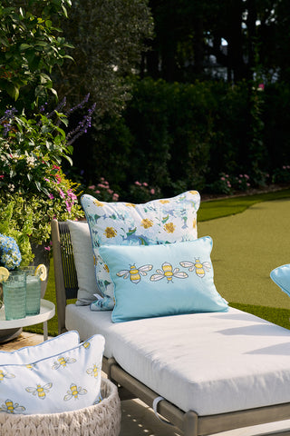 A sunlit outdoor scene features a white cushioned chaise lounge with Bee Trio Lumbar Outdoor Pillows. Nearby, a small side table holds a pitcher and glasses. The setting, surrounded by lush green foliage and flowers, benefits from durable SDS Shield® Fabric.