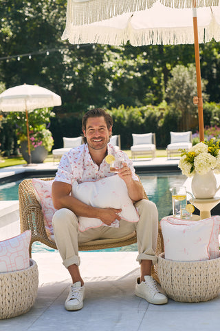 A person in a floral shirt and light pants sits on a wicker chair by the pool, holding a glass and the Flamingo with Pink Lattice Lumbar Outdoor Pillow. They smile brightly, surrounded by cushions and an umbrella, beautifully framed by the lush garden backdrop.