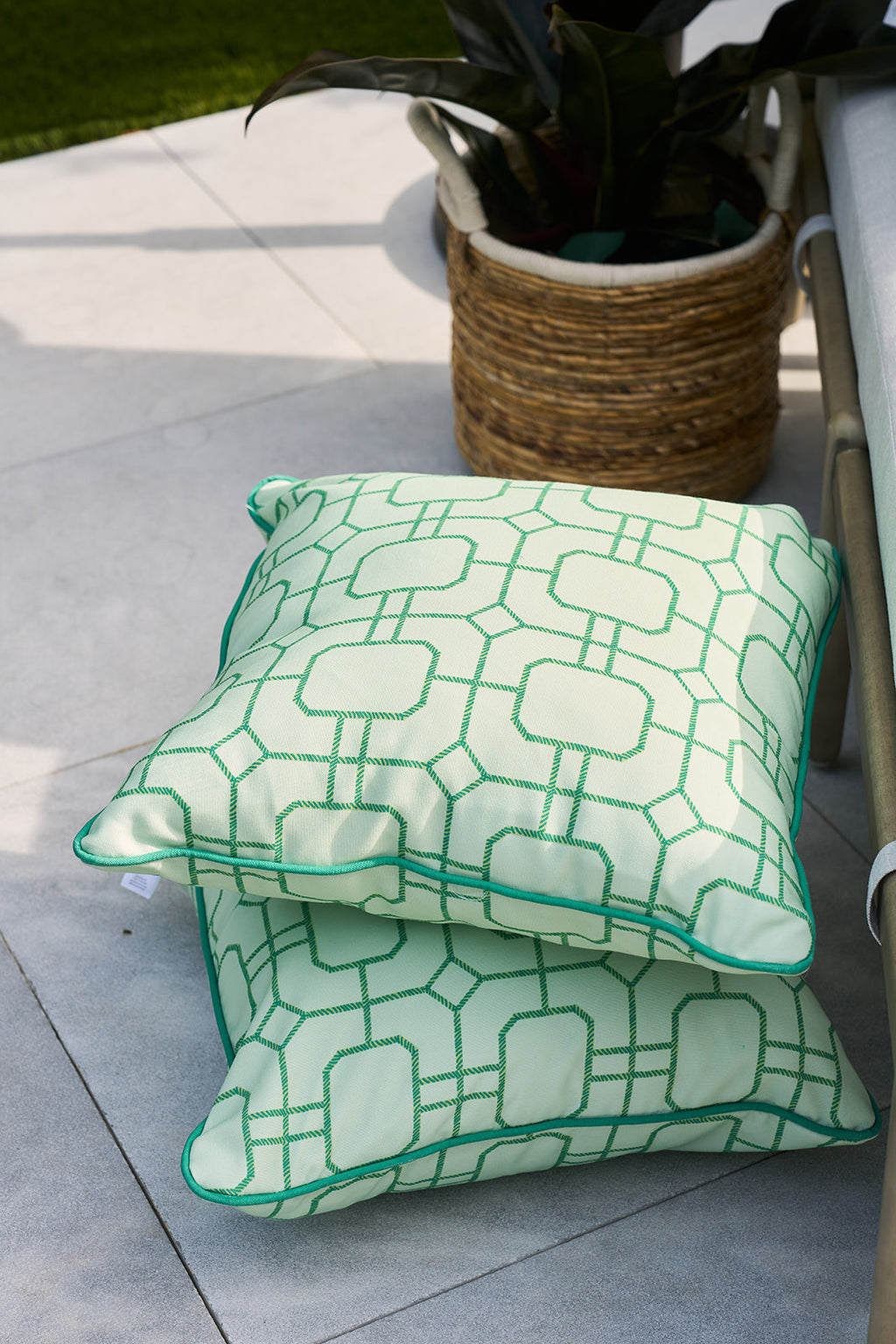 A person wearing a floral shirt holds a Green Lattice Outdoor Pillow from Sewing Down South®, featuring an elegant pattern of interlocking squares in vibrant green on a light background. The focus remains on the stylish pillow rather than their partially visible face.