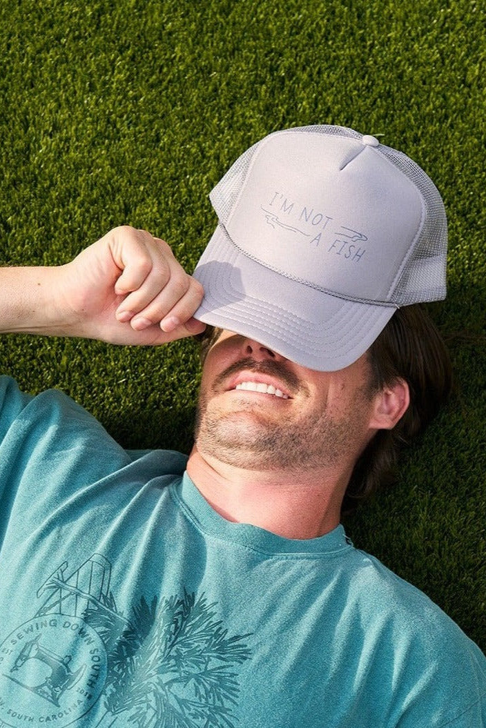 A light gray mesh-back trucker hat with IM NOT A FISH and a snorkel mask drawing on the front. This adjustable snapback cap, known as the Im Not A Fish Hat, is set against a pale stone surface with a dark, blurred background.