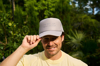 A man smiling outdoors adjusts his Im Not A Fish Hat. Hes wearing a light yellow T-shirt amidst lush greenery and trees under bright sunlight.