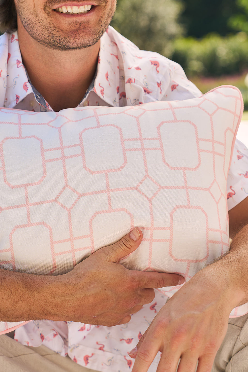 Sitting outdoors in a light shirt and pink shorts, a person holds the Flamingo with Pink Lattice Lumbar Outdoor Pillow. The serene scene features blurred greenery and part of another outdoor fabric pillow beside them.