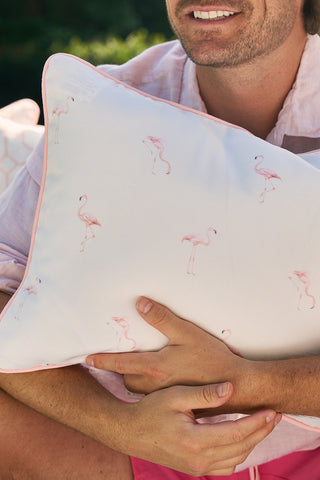 Sitting outdoors in a light shirt and pink shorts, a person holds the Flamingo with Pink Lattice Lumbar Outdoor Pillow. The serene scene features blurred greenery and part of another outdoor fabric pillow beside them.