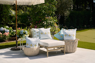 In a tranquil garden, the outdoor scene features a Sewing Down South Bee Trio with Repeating Bees Outdoor Pillow on a lounge chair. Wicker baskets and decorative pillows of durable outdoor fabric enhance the fringed umbrella, all amidst lush greenery and vibrant flowers.