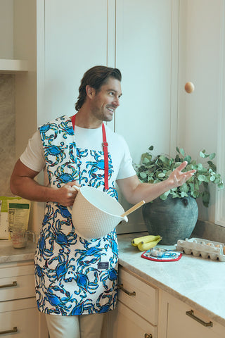 A man in a Crab Craze Apron with perfect pockets and adjustable neck stands playful in the kitchen with a mixing bowl and an egg. The counter features bananas, eggs, and a potted plant, all set against exclusive Sewing Down South patterns.