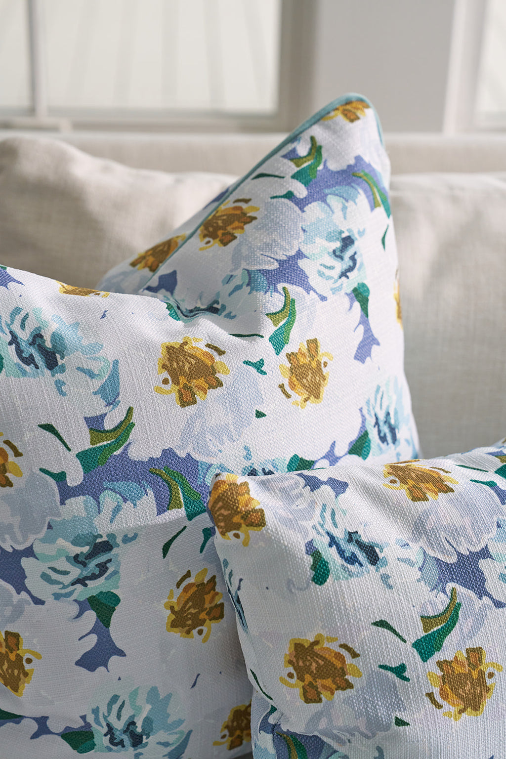 A person sits on a couch, holding a Blossoming Peonies Pillow from the Bee & Florals Collection. The sofa is decorated with complementary blue and yellow floral pillows by Sewing Down South®. A woven basket and potted plant enhance the bright, sunlit rooms charm.
