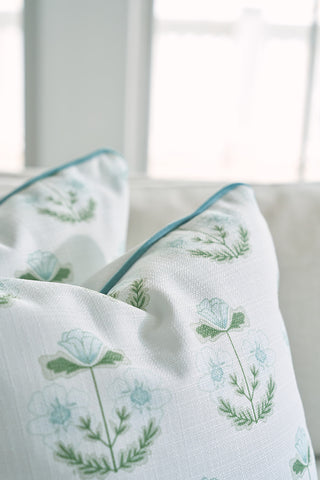 A close-up of two decorative pillows on a couch displays the Bee & Florals Collection: Victorian Floral in Blue Pillow, featuring green and light blue flowers. The airy background, softened by blurred windows, bathes the scene in gentle natural light.