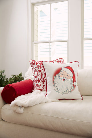 A white sofa with a Christmas Throw Pillow, a red bolster, fluffy white throw, and two festive pillows—a Handwritten Ho Ho Ho Pillow and a Vintage Stitch Santa Pillow—sits by large windows letting in natural light.