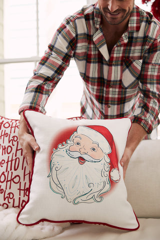 A smiling person in a plaid shirt holds a Vintage Stitch Santa Pillow featuring a cheerful Santa illustration, with other festive “Ho Ho Ho” pillows in the background.