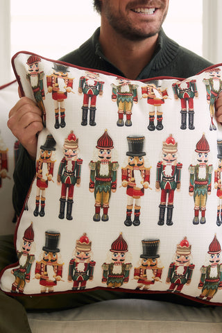 A person holds the Nutcracker Line Up Pillow, featuring colorful soldiers in festive outfits on a white background. With their face partially out of frame, they smile, showing off this perfect holiday accent.
