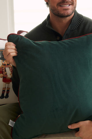 A man in a dark sweater smiles while holding a large green velvet pillow with red piping. In the background, the Nutcracker Line Up Pillow adds festive holiday charm to the scene.