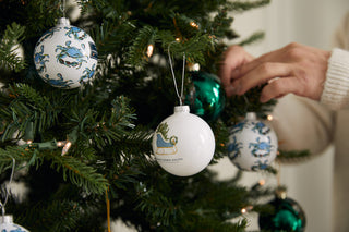 A person decorates a Christmas tree with white and green ornaments, including Sleigh Globe Ornaments (available in single, 2 or 4 packs) from Sewing Down South. The lush branches shimmer with sparkling lights and festive holiday decorations.