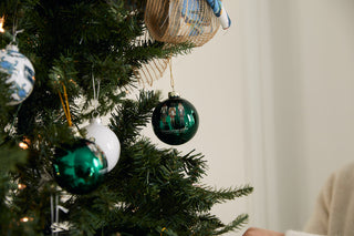 Close-up of a Christmas tree with green and white ornaments, including a shiny green bauble and Nutcracker Globe Ornaments (Single, 2 or 4 pack). A person in a cream sweater’s arm is partially visible on the right.