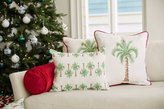 A beige sofa with the Holiday Palmetto Lumbar Pillow, palm tree-patterned pillows, and a red bolster sits next to a Christmas tree with white and teal ornaments in a bright room with large windows.