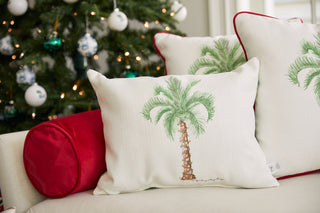 The Holiday Palmetto Lumbar Pillow, adorned with a palm tree and festive lights, rests on a white couch with a red bolster pillow. Behind it, a sparkling ornamented Christmas tree completes the cheerful holiday scene.