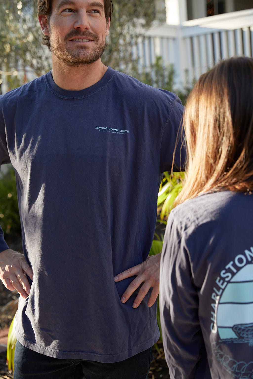 Two people walk down a sunny, palm-lined street, each wearing a Charleston Pineapple Long Sleeve Tee featuring Charleston, South Carolina and a Pineapple Fountain illustration on the back.