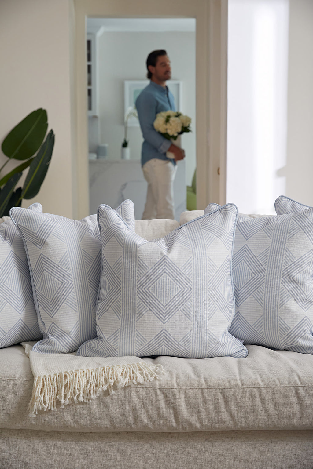 A man in a light blue shirt and white pants sits on a couch, smiling and holding the Oversized Mod Geo Diamond Pillow from Sewing Down South, which features a light blue and white geometric pattern.
