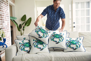 In a bright living room, someone arranges decorative pillows from the Coastal Collection on a white sofa. The King Crab Pillow with Navys vibrant design stands out amid a brick wall, potted plant, and glass door, enhancing the rooms coastal charm.
